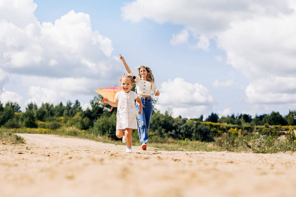 Ein lachendes kleines Mädchen und ihre Mutter laufen entlang der Straße zum Feld und lassen einen Drachen steigen. Mutter und Tochter haben gemeinsam Spaß - Foto, Bild