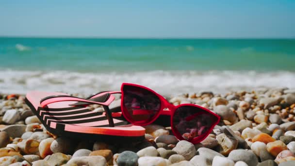 Zonnebrillen in de vorm van harten liggen op een kiezelstrand met zeegolven op de achtergrond. Het concept van een geweldige vakantie. 4k video. - Video