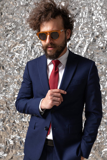 ελκυστικός επιχειρηματίας κρατώντας το ένα χέρι στην τσέπη ενώ προσαρμόζει το σακάκι του και φοράει γυαλιά ηλίου - Φωτογραφία, εικόνα