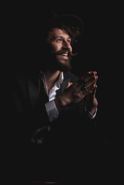 handsome businessman with a smile on his face is holding hands together and looking away on black background - Foto, imagen