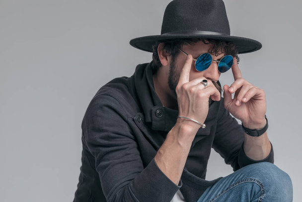 hűvös hipster hozzáállás rögzíti a napszemüveg, visel egy kalap és guggoló szürke háttér - Fotó, kép