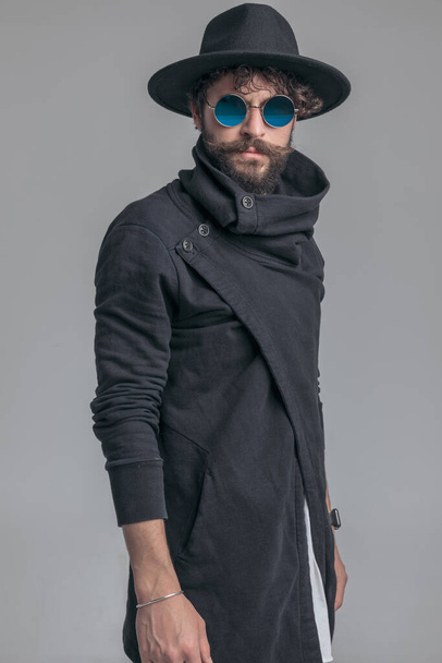 sexy uomo casual in posa con atteggiamento fresco e indossa un cappello, occhiali da sole e cardigan su sfondo grigio - Foto, immagini