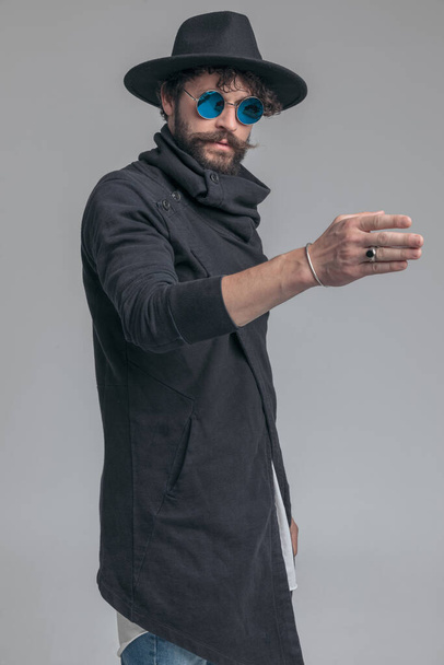 sexy casual man posing with one arm raised, wearing sunglasses, hat and a cardigan on gray background - Фото, зображення