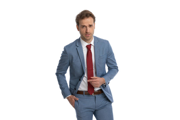Porträt eines sexy jungen Geschäftsmannes im blauen Anzug, der die Hand in der Tasche hält und auf modische Weise posiert, während er isoliert auf weißem Hintergrund im Studio steht - Foto, Bild