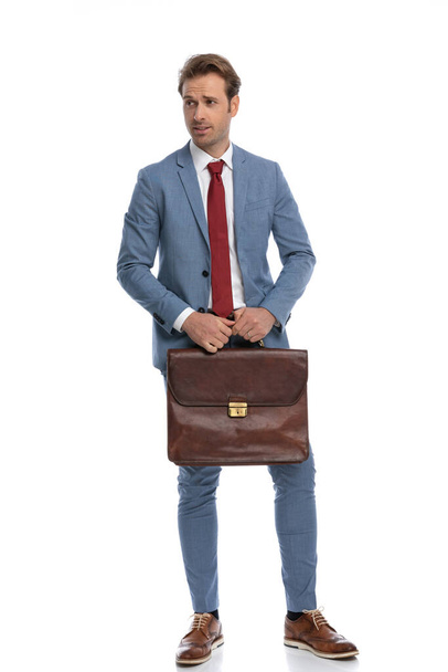 full body picture of sexy businessman in blue suit holding suitcase and looking to side while standing and posing on white background in studio - Foto, immagini