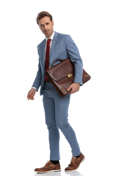 full body picture of sexy elegant man in blue suit walking while holding suitcase in front of white background in studio - Photo, Image