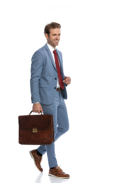 side view of elegant businessman in blue suit smiling while holding suitcase and walking isolated on white background in studio, full body - Foto, Bild