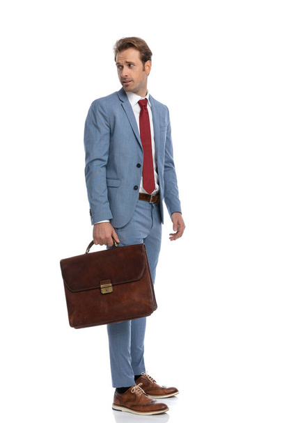 青いスーツケースを着た好奇心旺盛な若いビジネスマンが肩越しにスーツケースを見ながらスタジオの白い背景に並んで - 写真・画像