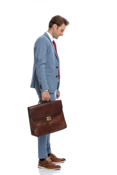 vista lateral del joven elegante en traje azul con la maleta de pie en la línea y mirando hacia abajo sobre fondo blanco en el estudio - Foto, imagen