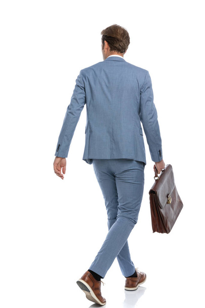 vista trasera del joven hombre de negocios en traje azul sosteniendo la maleta, mirando hacia un lado y caminando aislado sobre fondo blanco en el estudio - Foto, Imagen