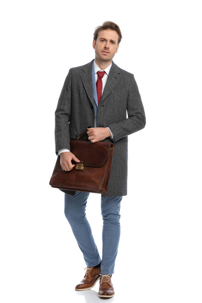 full body picture of elegant man in suit with jacket holding and opening briefcase while walking isolated on white background in studio - Zdjęcie, obraz