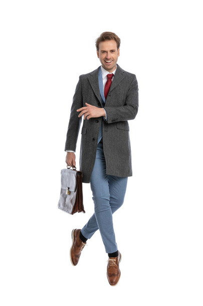 full body picture of happy young businessman with suitcase jumping in the air and laughing on white background in studio - Photo, Image