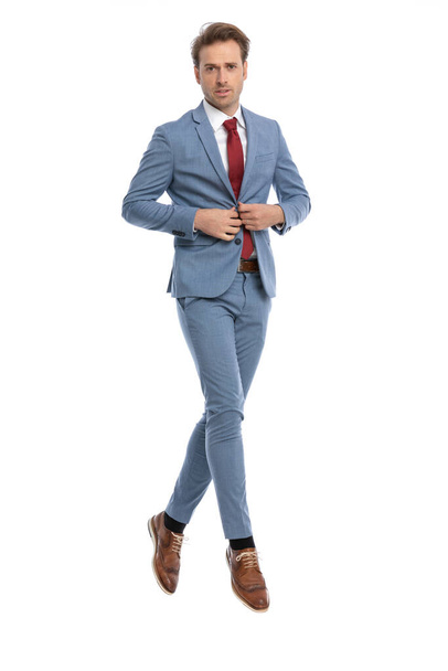 handsome elegant businessman buttoning blue suit and jumping in the air on white background in studio, full body - Zdjęcie, obraz