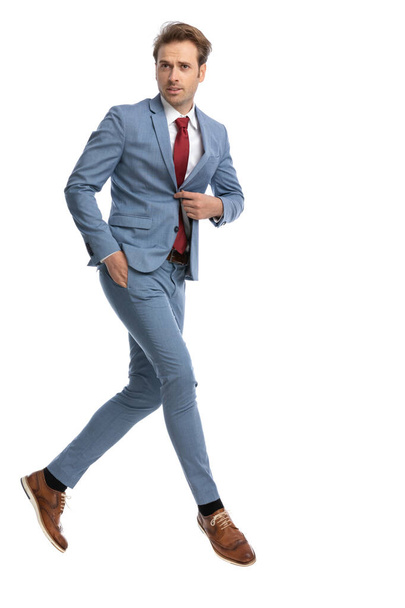 cool young guy holding hand in pocket and unbuttoning blue suit while jumping in the air on white background in studio, full body - Foto, Imagen