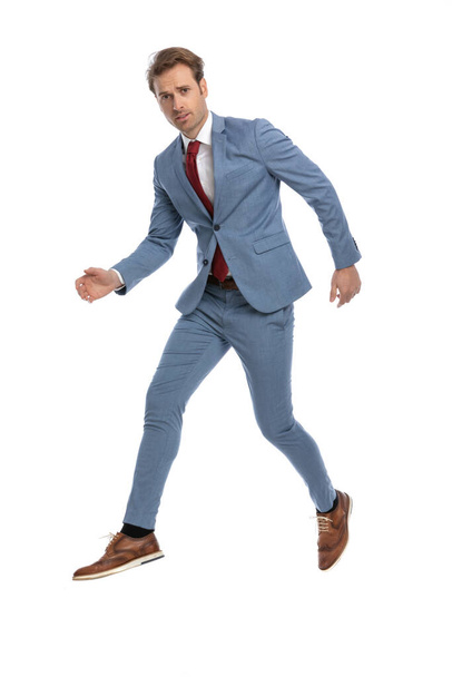 elegant young businessman in blue suit holding arms in fashion pose and jumping in the air on white background in studio, full body - Foto, afbeelding