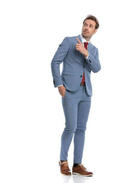 привлекательный молодой человек в синем костюме держит руку в кармане, указывая пальцем вверх и в сторону и ходьба изолированы на белом фоне в студии - Фото, изображение