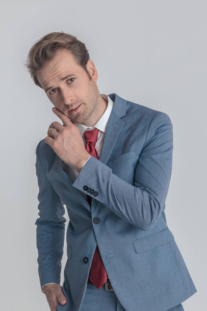 eleganter junger Mann im Bolue-Anzug hält Hand in Hand in einer Mode-Pose und berührt Gesicht, steht auf grauem Hintergrund und posiert im Studio, Porträt - Foto, Bild
