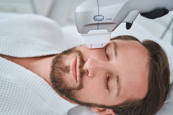 Bearded mustached dark-haired man dozing during a non-invasive face-lift procedure - Photo, Image