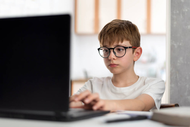 Child sits at table with laptop and does homework. Teen boy with glasses is studying online. Distance learning. - Photo, Image