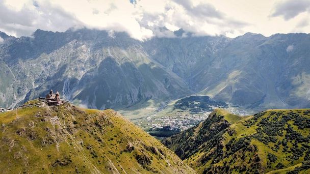 Drohnenaufnahme der Natur in Georgien. Kaukasus-Gebirge, Gergeti-Dreifaltigkeitskirche auf einem Hügel, Grün, Tal, Dorf im Hintergrund - Foto, Bild