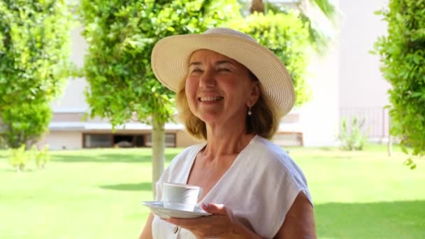 dromer Een volwassen vrouw van 50-55 jaar in een strohoed drinkt 's morgens vroeg op het terras van haar huis in een groene tuin op een zonnige dag een warme borrel koffie of thee uit een wit kopje. Senior senior - Video