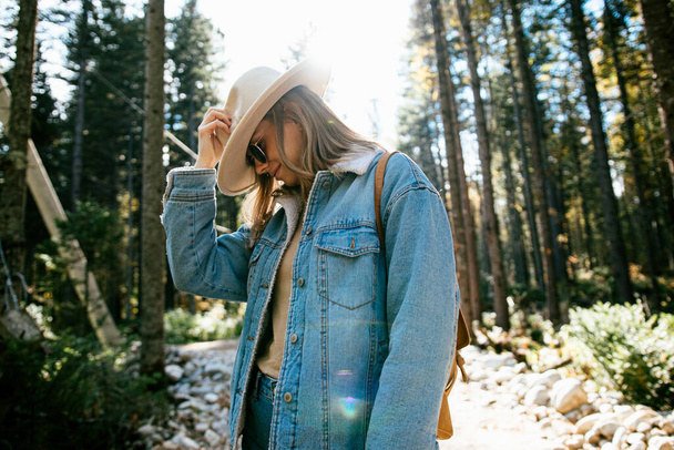 流行の帽子とデニムのスーツにサングラスとバックパックを身に着けて美しい女の子は秋の森を歩く - 写真・画像