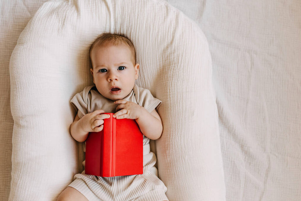 Bebé acostado en un nido de bebé, con un libro en blanco con cubierta roja. - Foto, imagen
