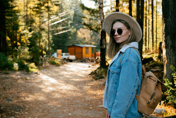 Güz ormanlarında gezen şık şapkalı, kot elbiseli, güneş gözlüklü güzel bir kız. - Fotoğraf, Görsel