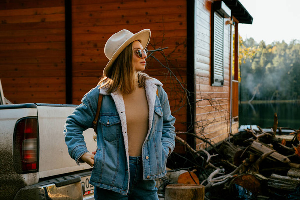menina bonita vestindo chapéu na moda e traje de ganga com óculos de sol posando perto de casa de campo de madeira e carro na floresta de outono - Foto, Imagem