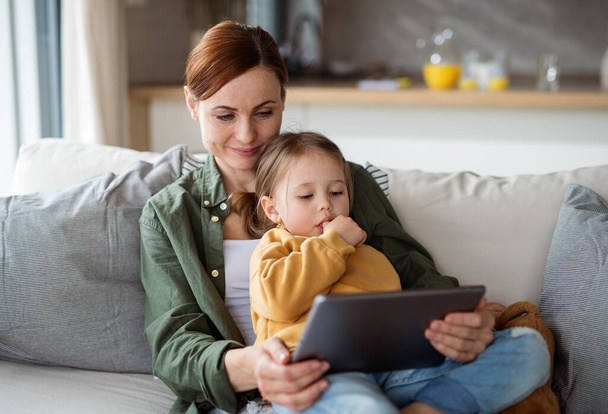 Happy mother with small daughter watching kids programme on tablet indoors at home, single parenting concept. - Фото, изображение