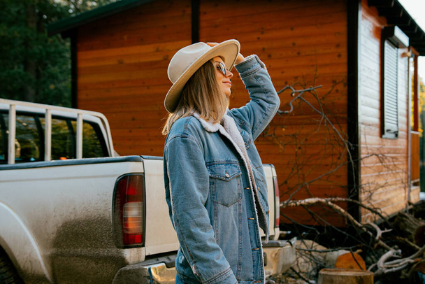 όμορφο κορίτσι φορώντας μοντέρνο καπέλο και denim κοστούμι με γυαλιά ηλίου ποζάρουν κοντά σε ξύλινο εξοχικό σπίτι και το αυτοκίνητο στο δάσος φθινόπωρο - Φωτογραφία, εικόνα