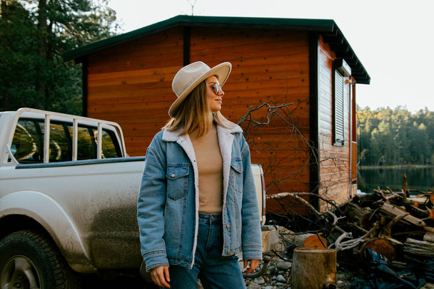 beautiful girl wearing trendy hat and denim suit with sunglasses posing near wooden country house and car in autumn forest - Fotoğraf, Görsel