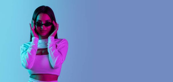 Cropped portrait of young beautiful girl posing in sunglasses isolated over blue background in neon light. Flyer - 写真・画像