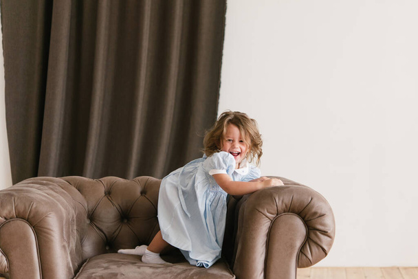 Little girl with a happy smile. Dressed in a blue dress. The girl sits on a brown armchair and indulges. - Photo, Image