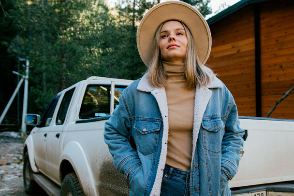 красивая девушка в модной шляпе и джинсовом костюме позирует возле деревянного загородного дома и автомобиля в осеннем лесу - Фото, изображение