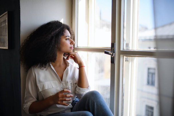 Portrait of young woman sitting on windowsill indoors, looking out and daydreaming. - Foto, Bild