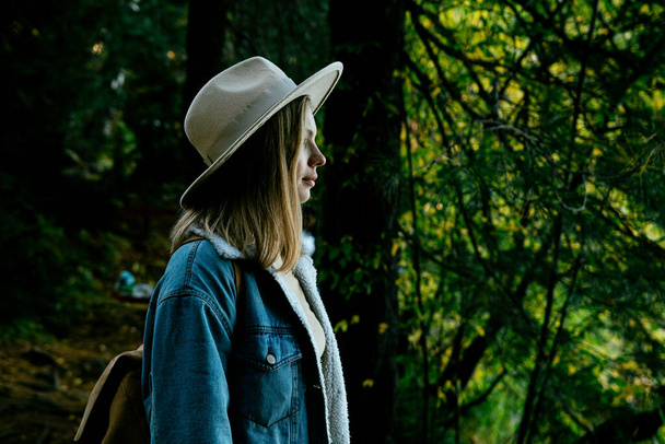 Güz ormanlarında gezen şık şapkalı, kot elbiseli, güneş gözlüklü güzel bir kız. - Fotoğraf, Görsel