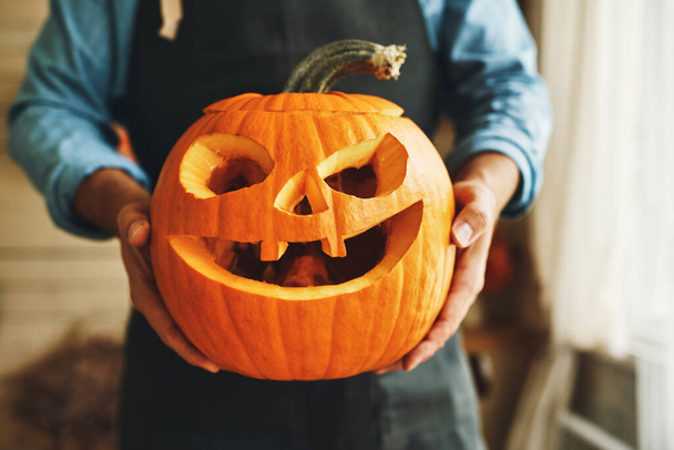 Cropped shot of man in apron with Halloween pumpkin monster in hands, person showing at camera classic carven spooky jack-o-lantern with big, gap-toothed grin, triangular eyes and nose - Φωτογραφία, εικόνα