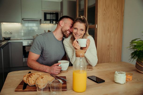 Καυκάσιο ευτυχισμένο ζευγάρι κάθεται στο τραπέζι της κουζίνας πίνοντας ζεστό καφέ  - Φωτογραφία, εικόνα