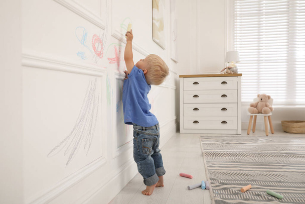 Mischievous little boy drawing with colorful chalk on white wall at home - 写真・画像