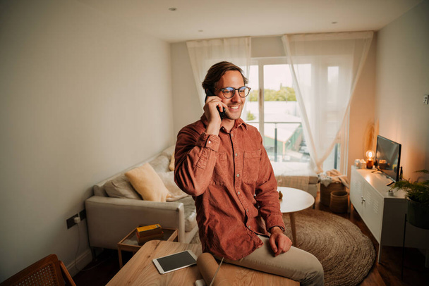 Καυκάσιος φοιτητής συνομιλίας σε κινητό τηλέφωνο τοποθέτηση σε γραφείο στο σπίτι συσταθεί - Φωτογραφία, εικόνα