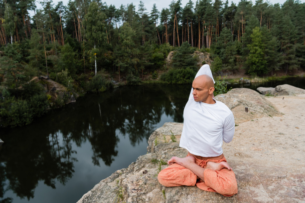 buddhist en sudadera blanca meditando en pose de loto con las manos detrás de la espalda en acantilado rocoso sobre el río - Foto, Imagen