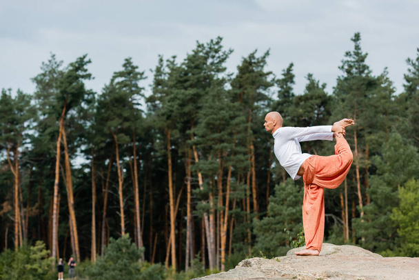 side view of buddhist practicing standing bow pose on rocky cliff in forest - Photo, Image