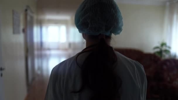 Uma médica cansada de roupas médicas caminha lentamente pelo corredor vazio de uma clínica médica ou hospital.  - Filmagem, Vídeo