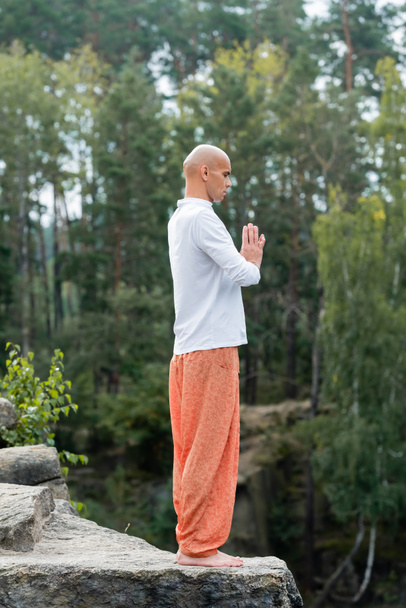 visão comprimento total do budista em sweatshirt e calças harém meditando com as mãos orando ao ar livre - Foto, Imagem