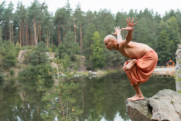 shirtless buddhist meditating in yoga pose with outstretched hands on rocky cliff over river - Fotó, kép
