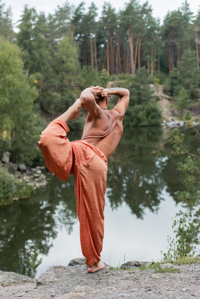 bouddhiste torse nu en pantalon de harem pratiquant une pose d'arc à une jambe sur une falaise rocheuse au-dessus d'un lac forestier - Photo, image