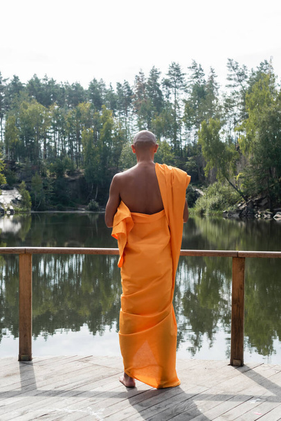 vue arrière du bouddhiste en kasaya orange méditant près du lac en forêt - Photo, image