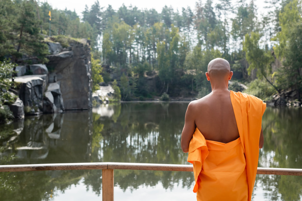 Вид сзади баддиста в оранжевой майке, сидящего возле лесного озера  - Фото, изображение