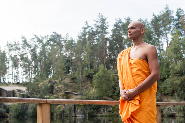 Буддийский монах в оранжевой касайе смотрит в сторону, медитируя возле деревянного забора в лесу - Фото, изображение